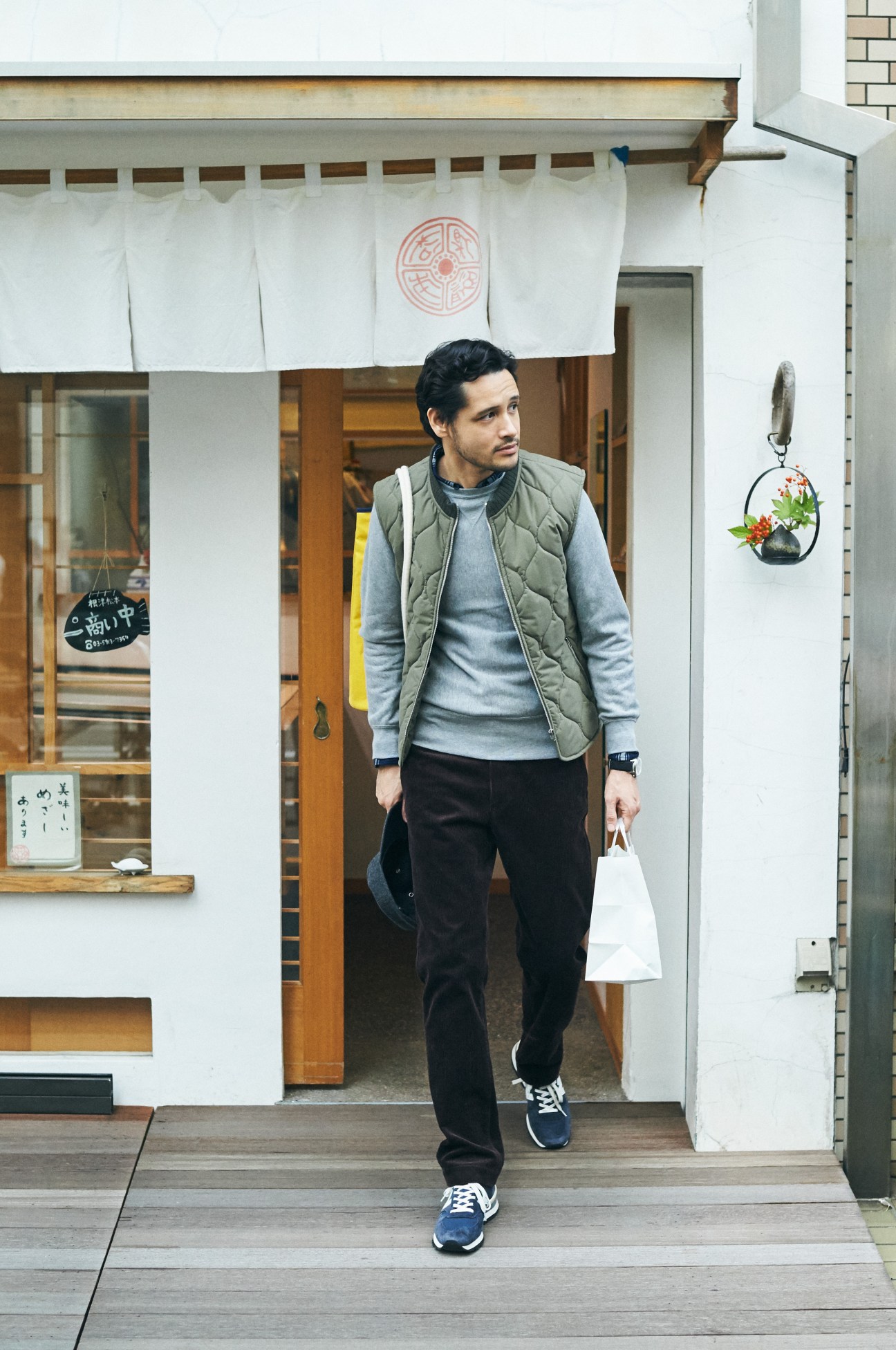 Man exiting a Japanese shop wearing a quilted down vest over a sweatshirt, holding a white shopping bag.