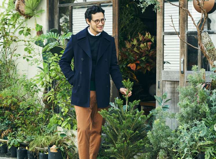 Man in navy coat and brown trousers standing outside a plant-filled storefront holding fresh vegetables.