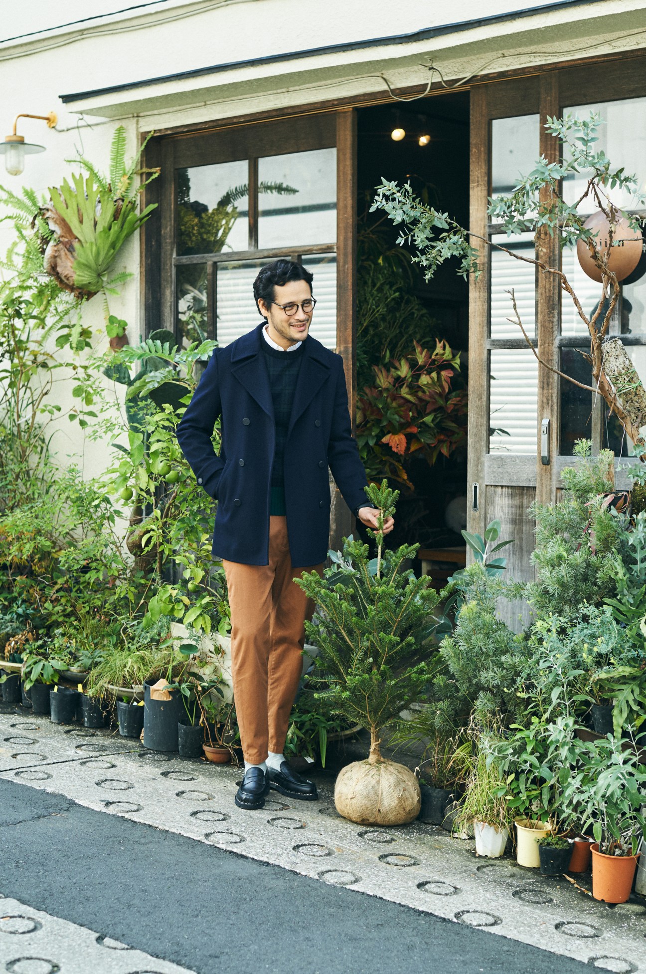 Man in navy coat and brown trousers standing outside a plant-filled storefront holding fresh vegetables.