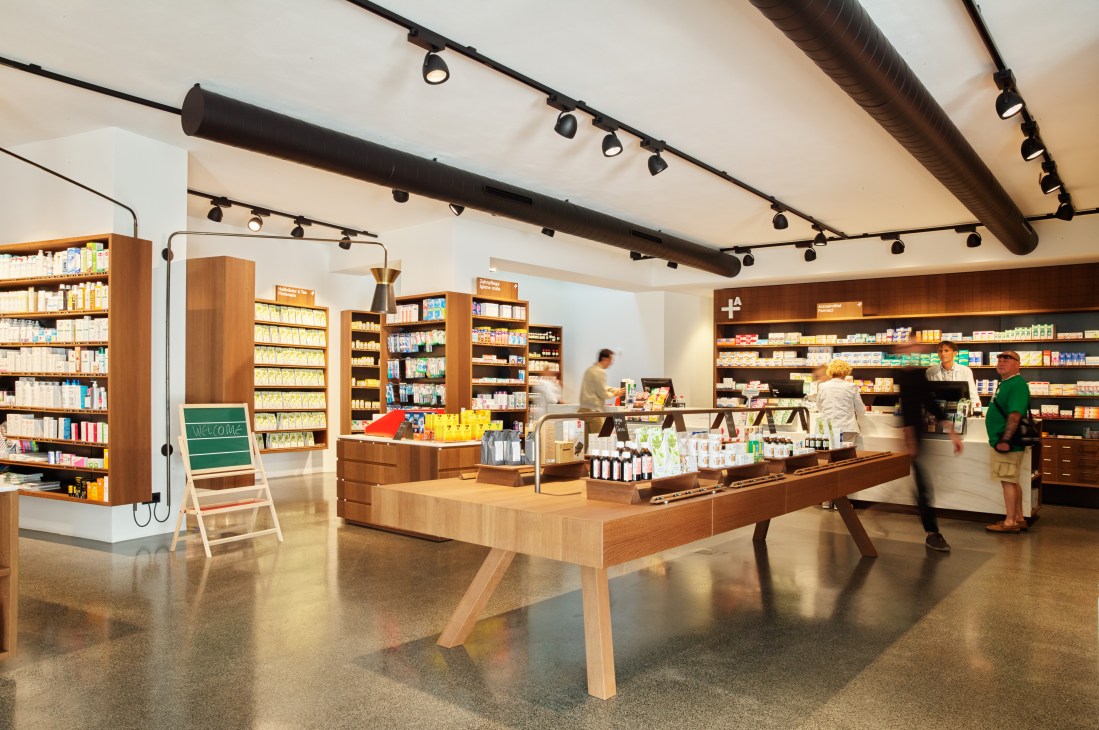 Modern pharmacy interior with wood shelving, track lighting, central display table, and customers browsing products