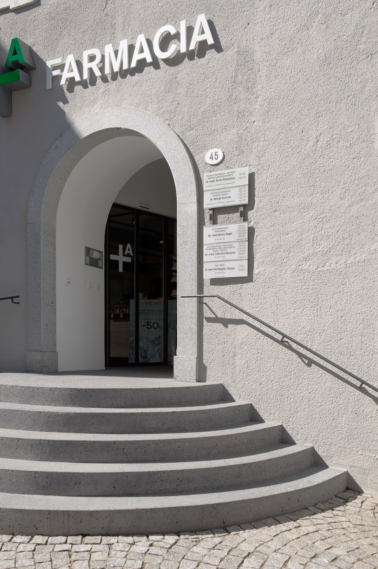 White building entrance with arched doorway, steps, and 'FARMACIA' sign with green cross above