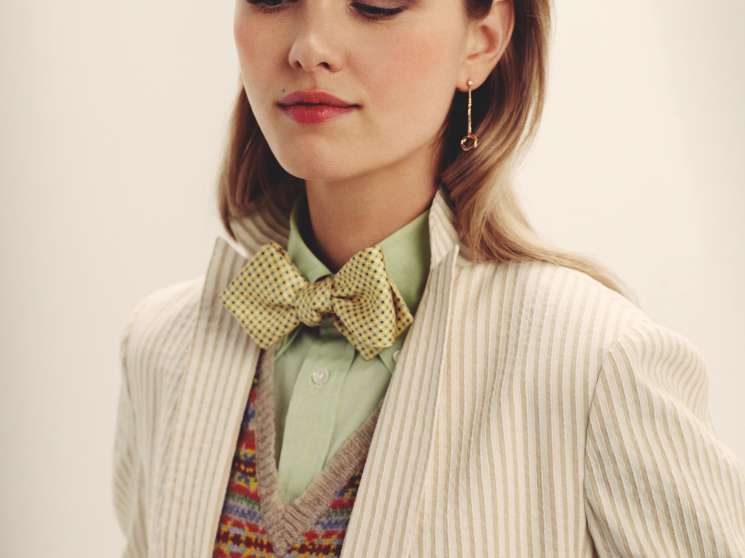Woman wearing cream pinstriped jacket, green shirt, patterned v-neck jumper and yellow bow tie with Cartier earrings.