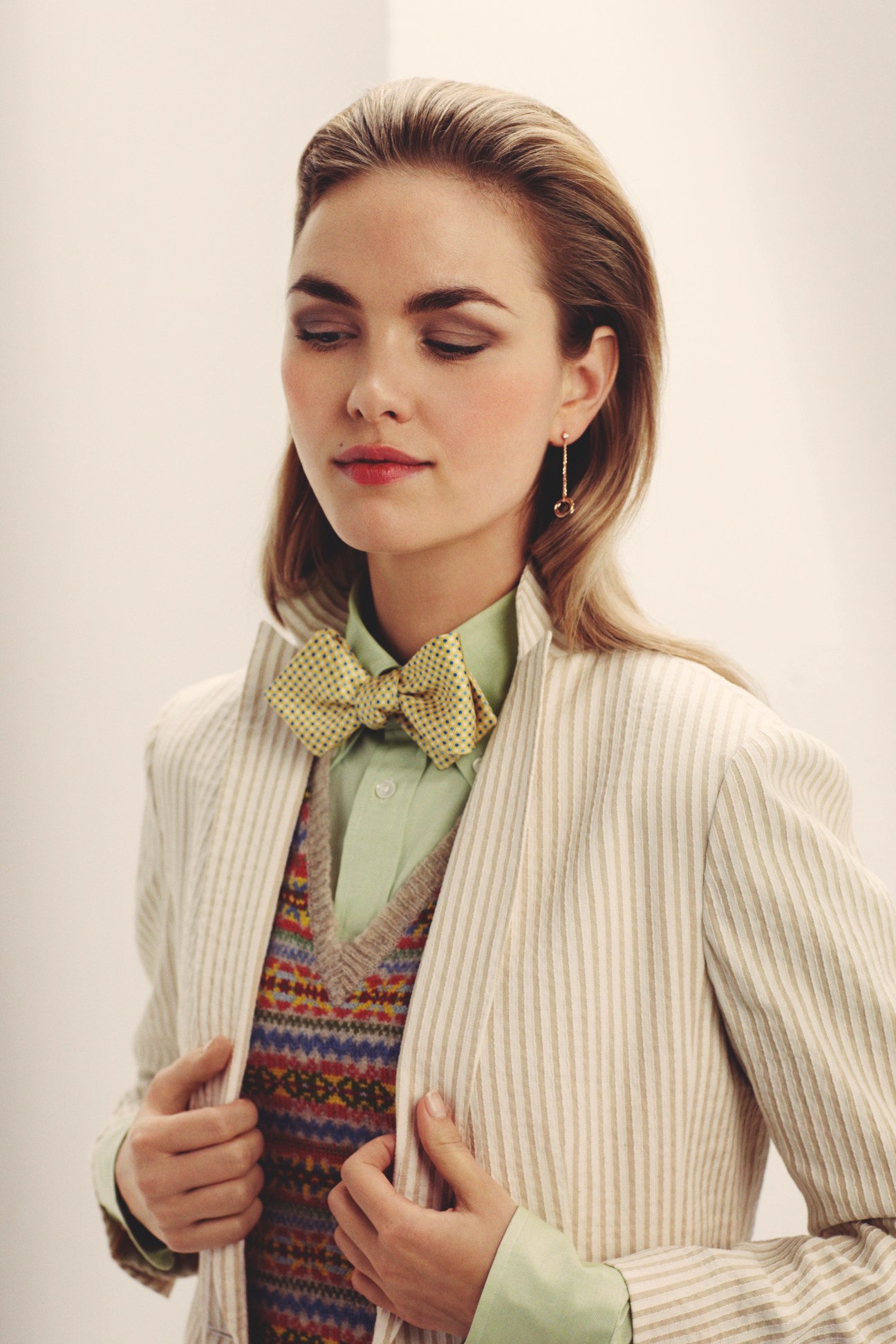 Woman wearing cream pinstriped jacket, green shirt, patterned v-neck jumper and yellow bow tie with Cartier earrings.