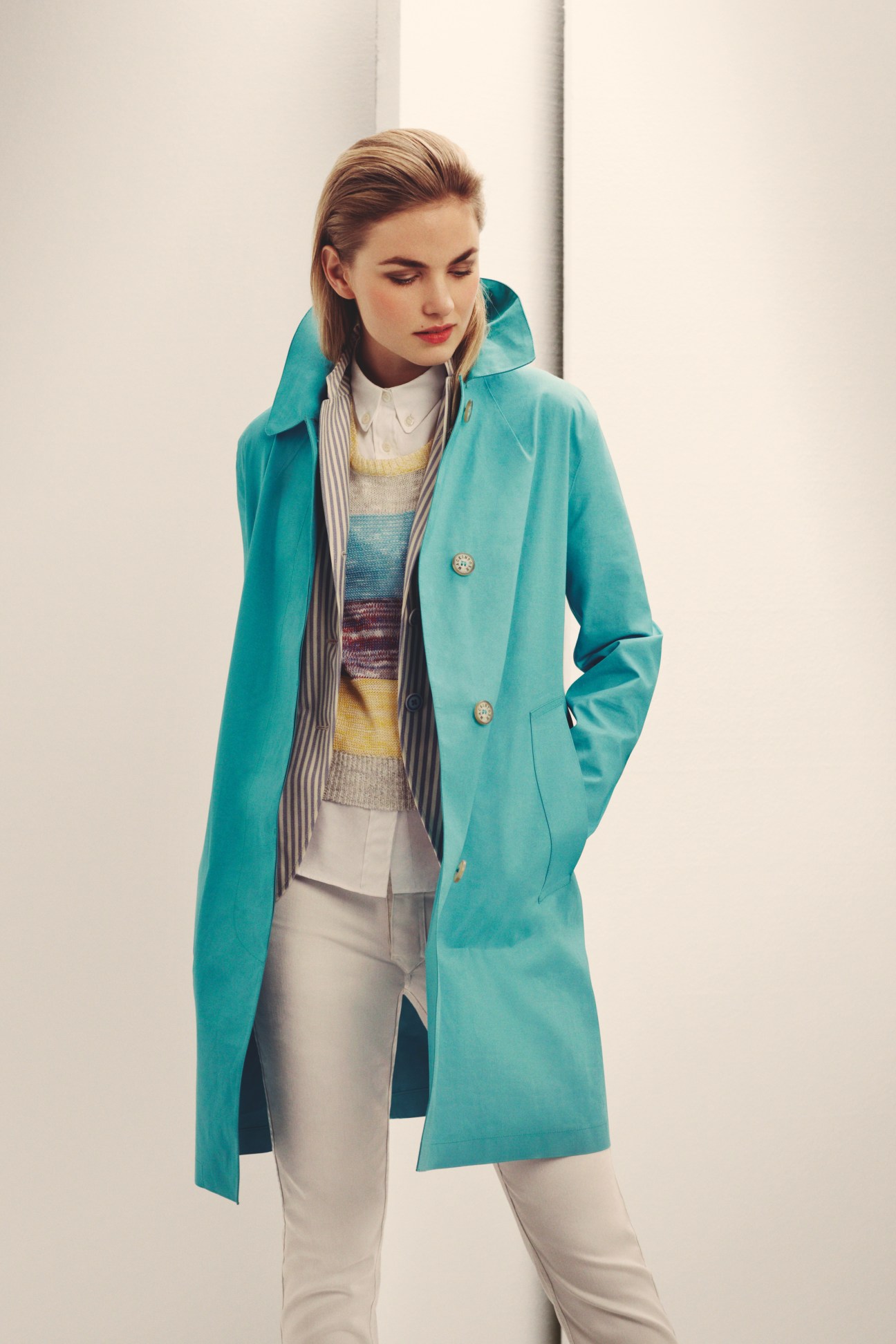 Woman wearing a turquoise Mackintosh coat layered over a striped shirt and colorful jumper with white trousers.
