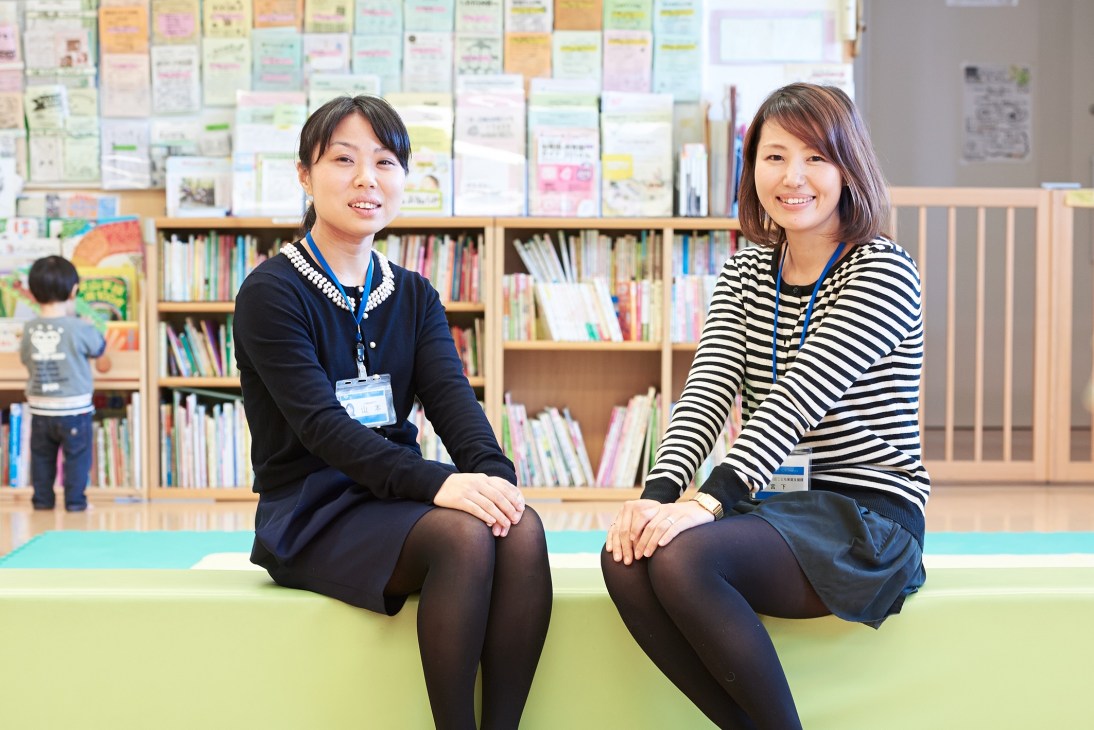 Two women wearing lanyards sit in a library with children's books and colorful drawings on the wall behind them.