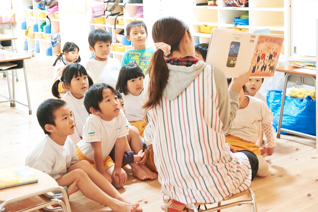 A caregiver reads a picture book to a group of young children sitting on the floor in a bright nursery classroom.