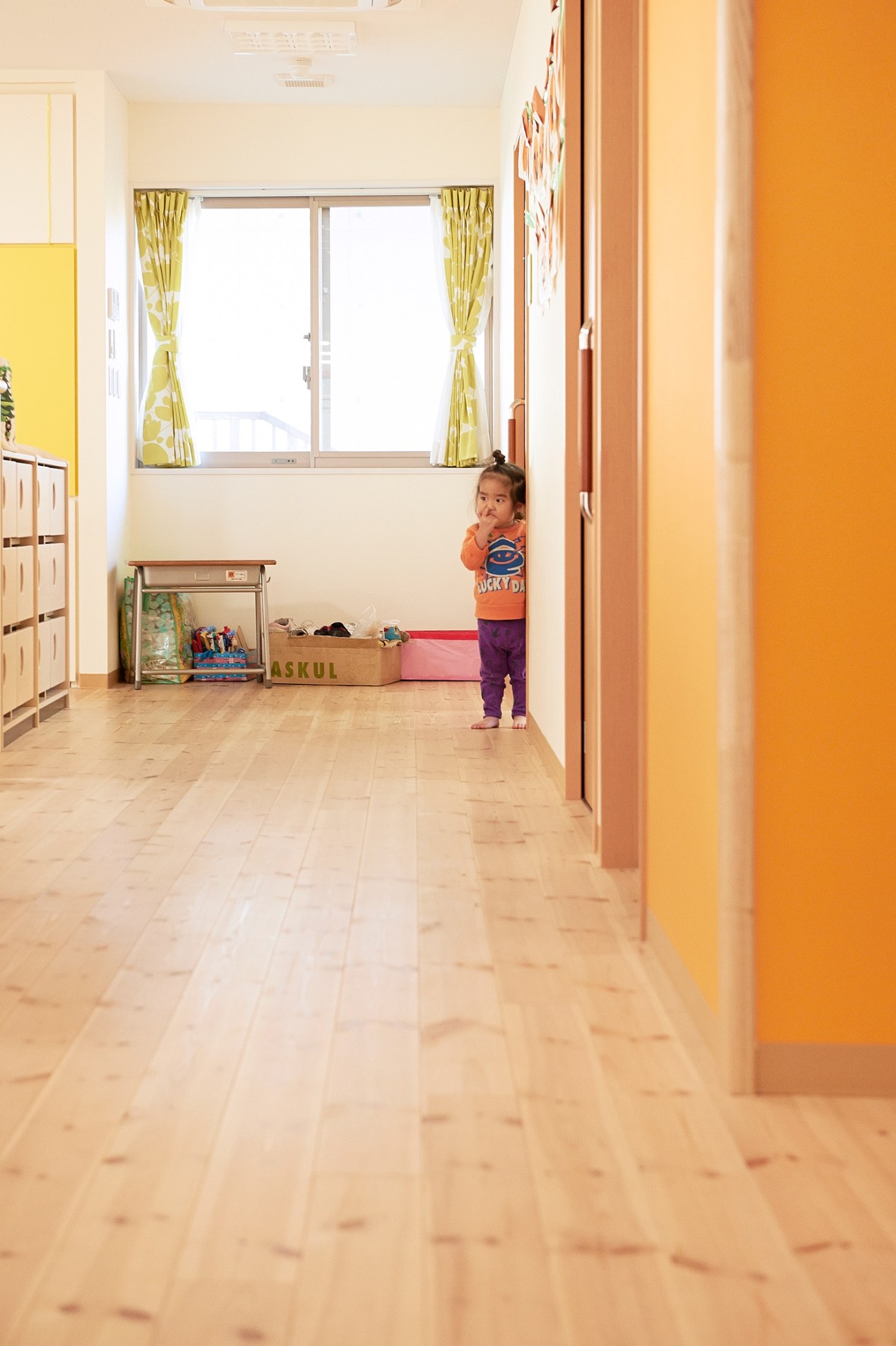 A young child stands in a bright, minimalist classroom with wooden floors, yellow walls, and large windows with yellow curtains.