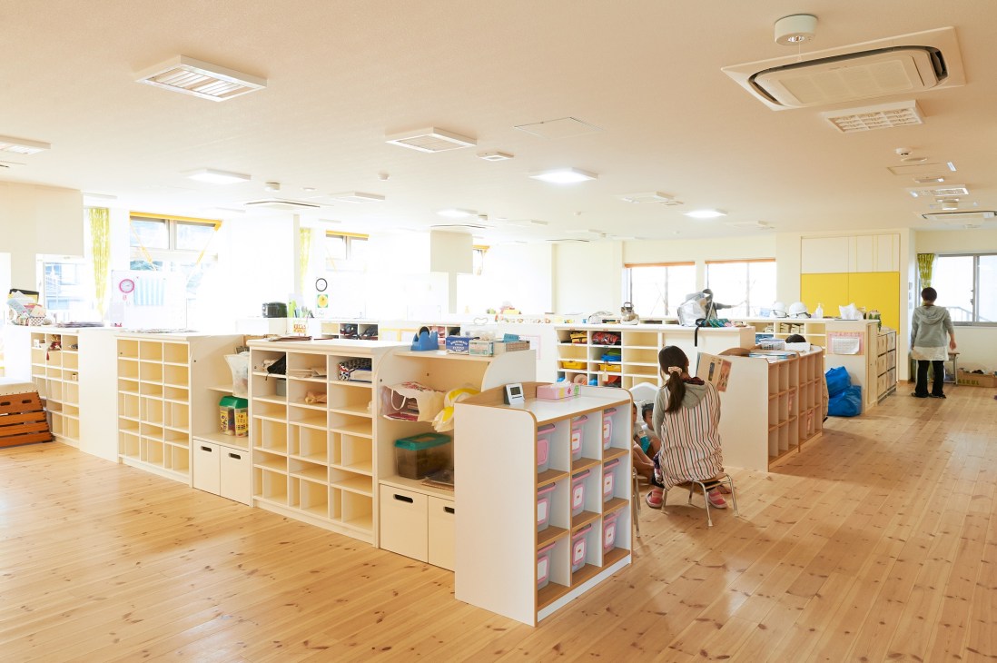 Spacious nursery classroom with wooden floors, natural light, and open shelving units storing toys and supplies.