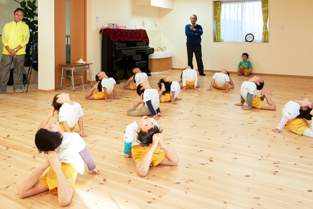 Young children in matching yellow and white outfits perform gymnastics exercises on a wooden floor at Byobugaura Harukaze Nursery in Yokohama, with instructors supervising.