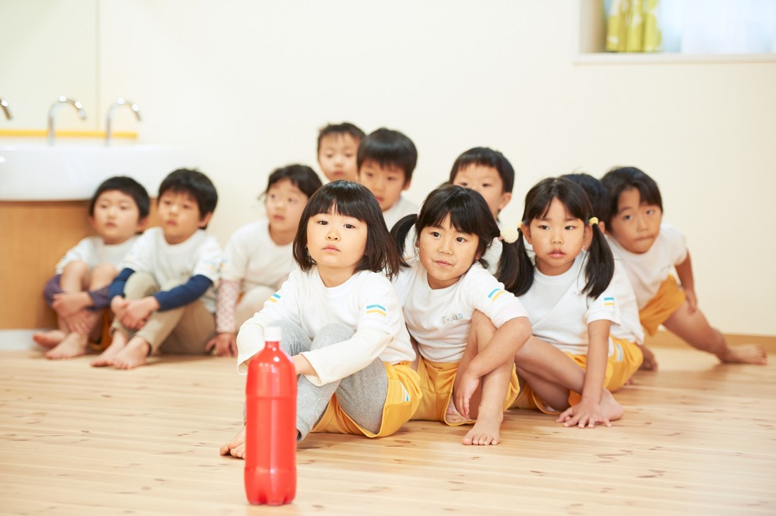 Young children sitting in a line on a wooden floor wearing white and yellow uniforms in a nursery classroom.