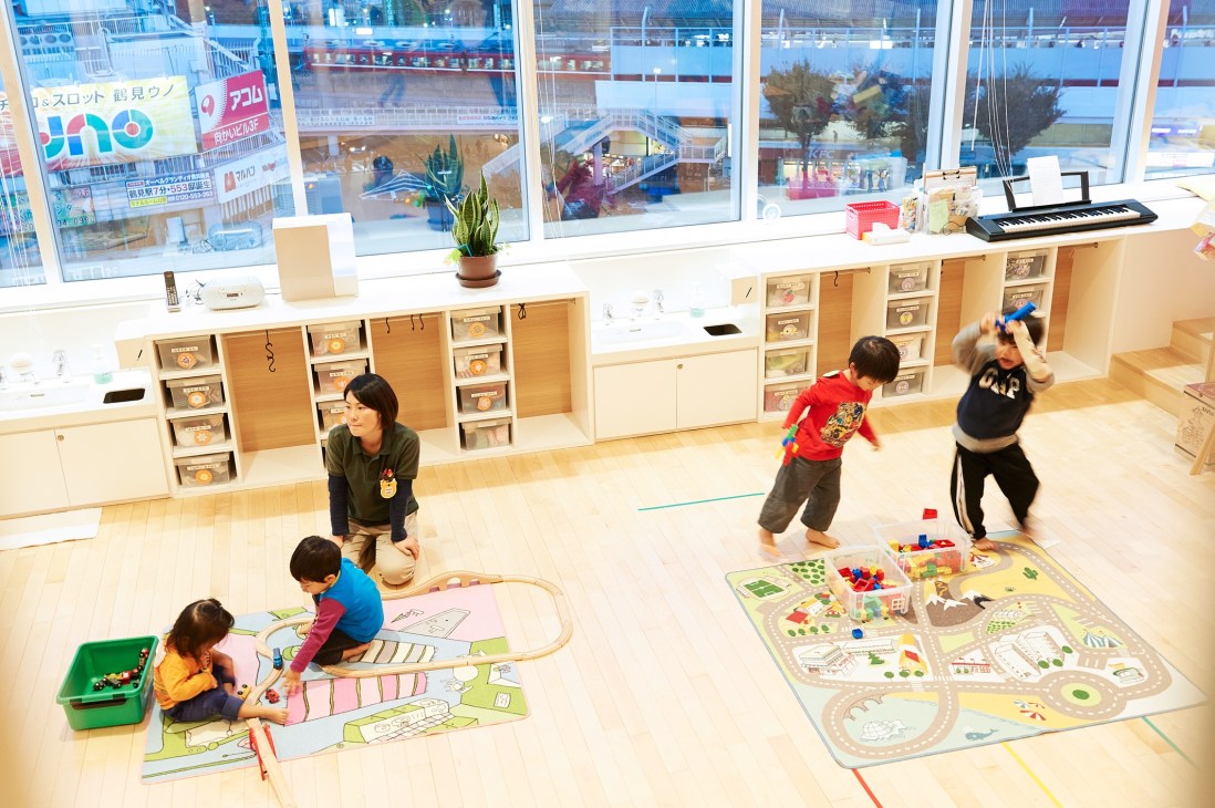 Children and a caregiver play in a bright, modern nursery classroom with wooden storage units and large windows overlooking the city.