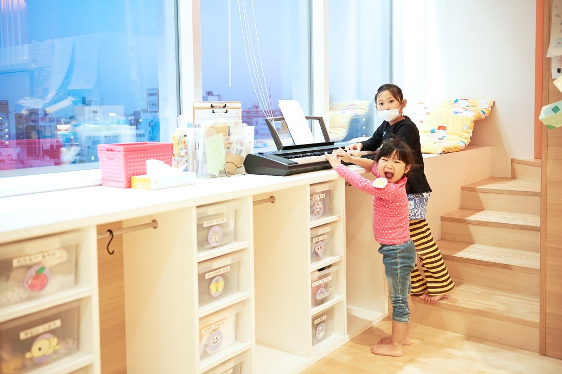 Two young children play a keyboard in a bright nursery room with city views and wooden furnishings at P's Smile.