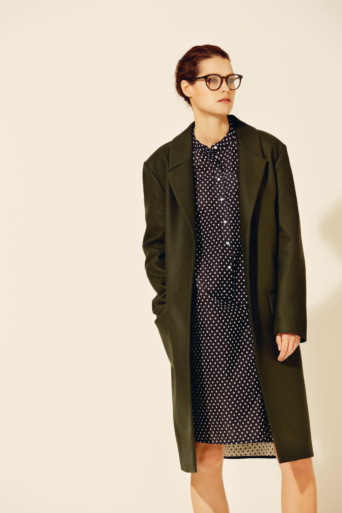 Woman wearing olive green wool coat over polka dot blouse with tortoiseshell-frame glasses.