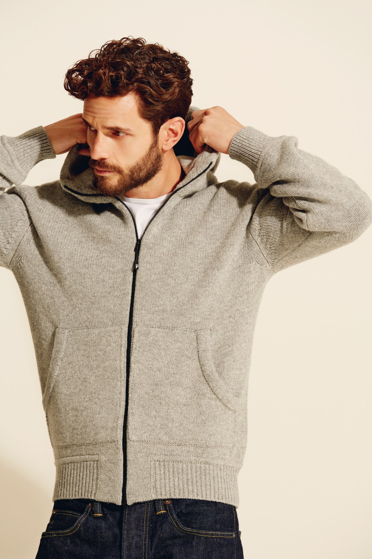 Man wearing a beige zip-up hooded wool cardigan with dark jeans against a neutral background.