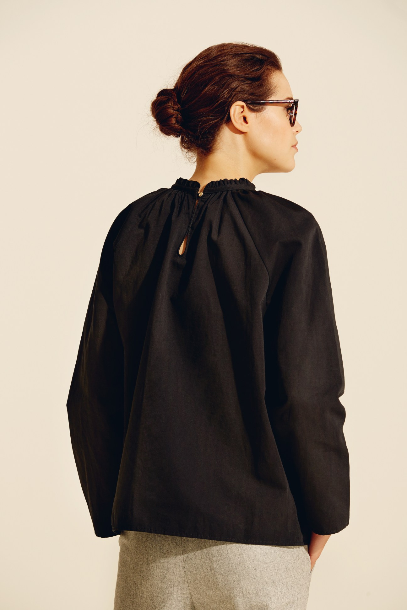Woman wearing black long-sleeved blouse with gathered neckline and beige trousers, viewed from behind.