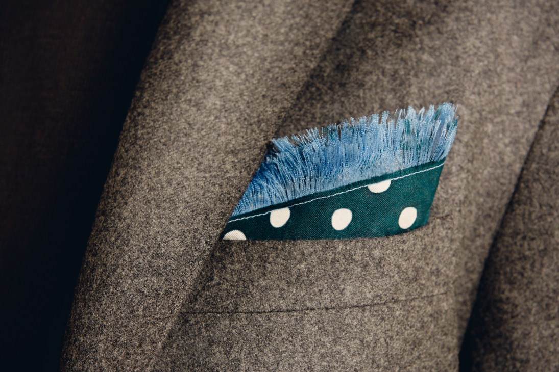 A fringed green-and-azure silk pocket square pinned to a grey wool jacket.