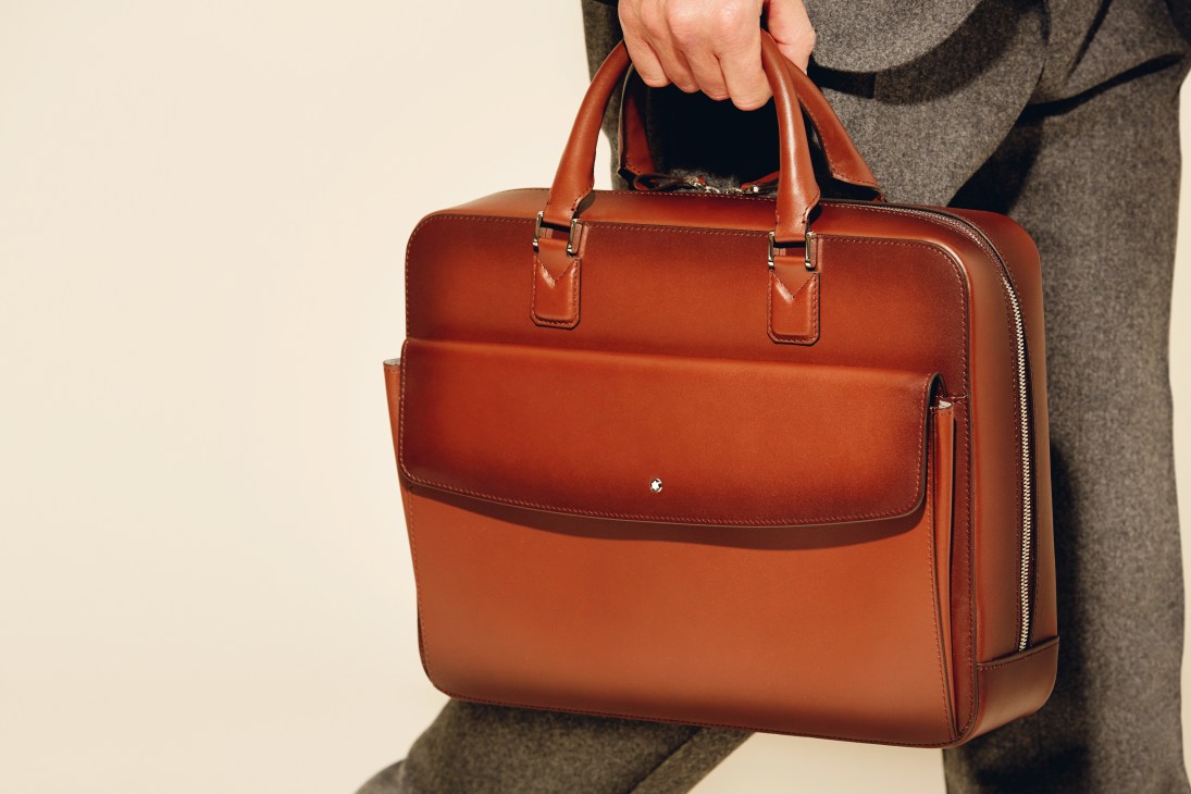 A person wearing a grey coat holds a cognac leather briefcase with tan handles and a front flap pocket.
