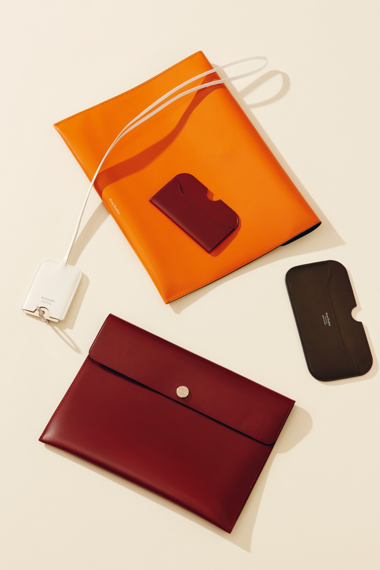 Acne leather accessories including an orange tote bag, burgundy wallet, burgundy phone holder and black case on a cream background.