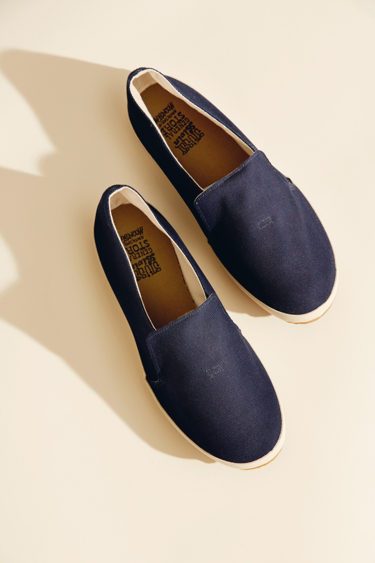 Pair of navy canvas slip-on shoes with white trim and cream-colored soles on a beige background.