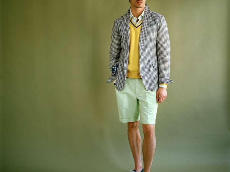Male model wearing yellow jumper, checkered blazer, and pale green shorts against green background.