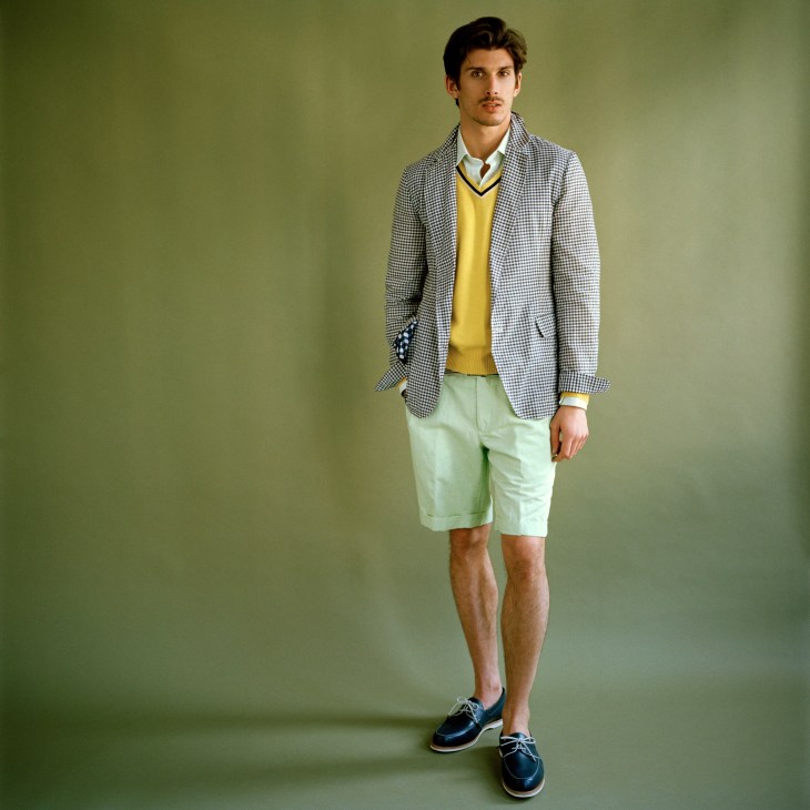 Male model wearing yellow jumper, checkered blazer, and pale green shorts against green background.