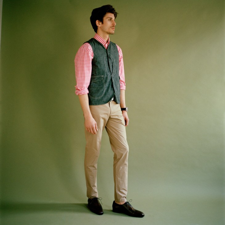 Male model wearing Post Overalls waistcoat, Hackett shirt, Aquascutum trousers, Berluti shoes and Trussardi 1911 belt against green background.