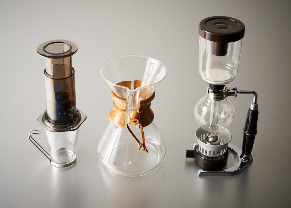 Three coffee makers displayed left to right: AeroPress, Chemex with wooden collar and leather tie, and Hario siphon brewer.
