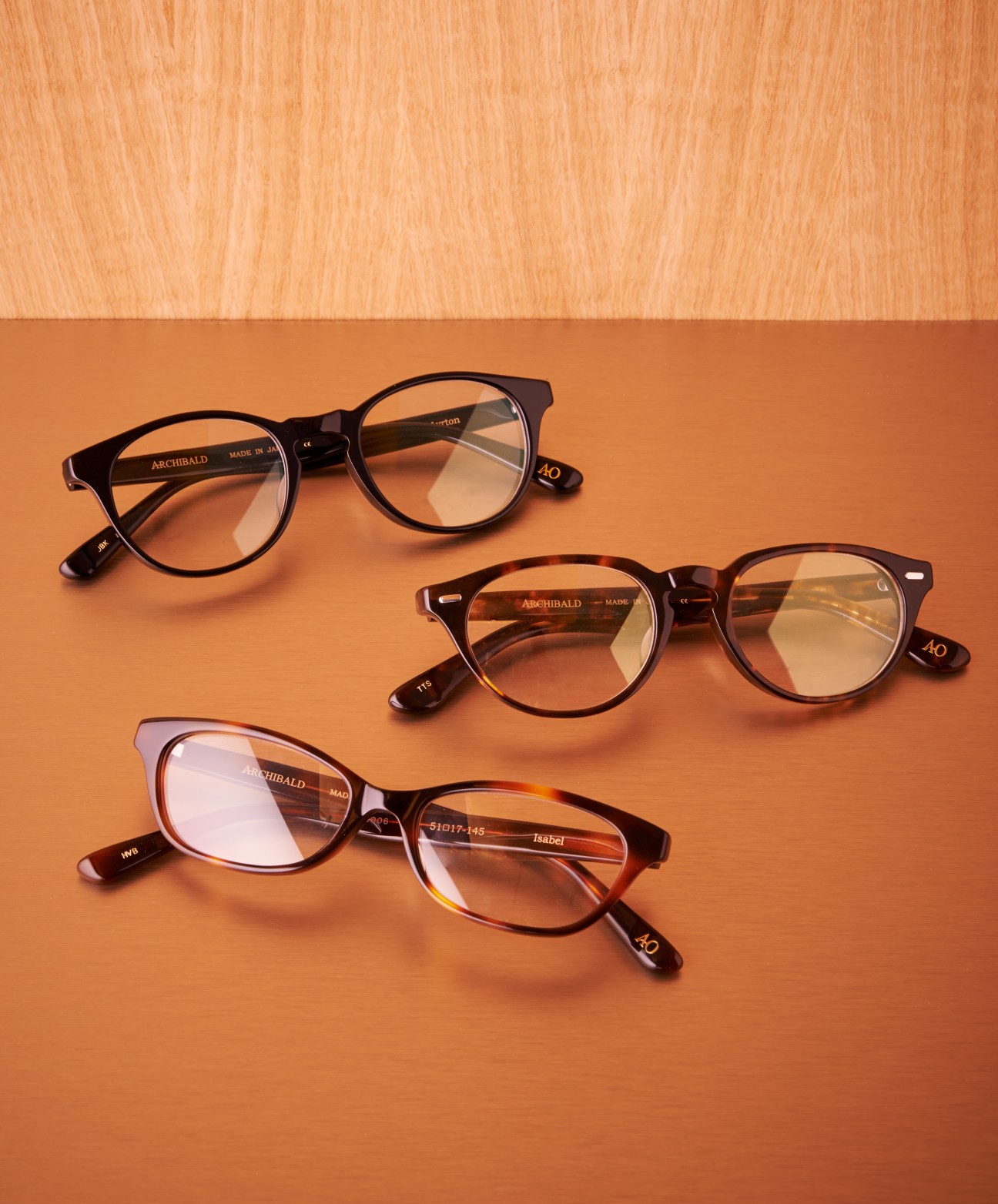 Three pairs of Archibald Optics glasses in black and tortoiseshell acetate frames displayed on an orange background.