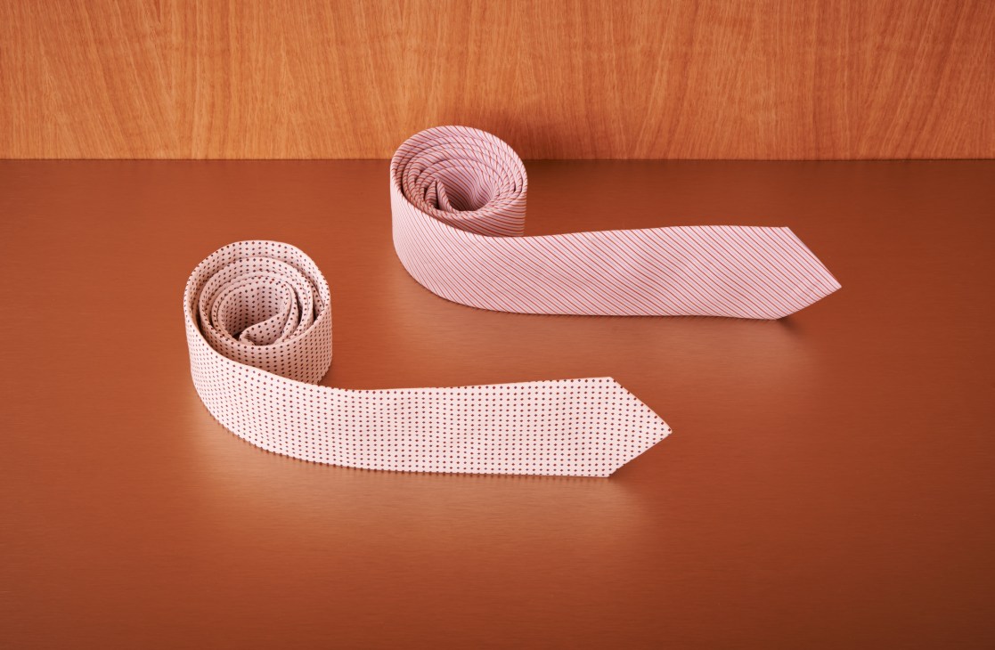 Two rolled neckties displayed on a terracotta background, one with a dotted pattern and one with diagonal stripes in pink tones.