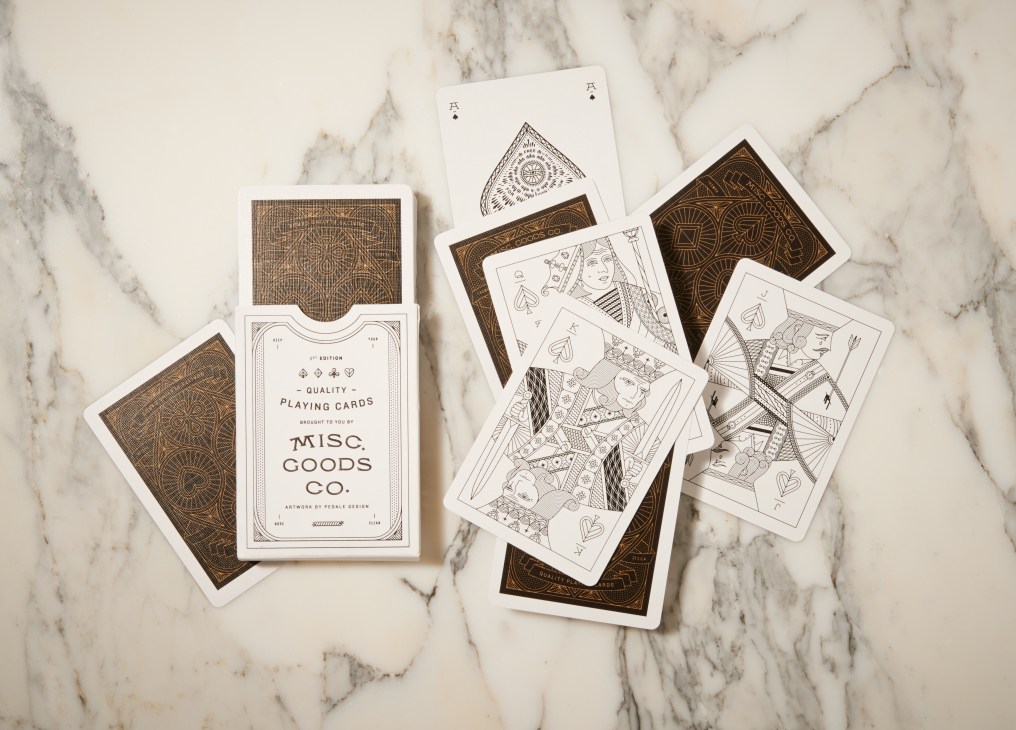 Misc. Goods Co. custom-designed playing cards with gold and white deck designs on marble surface.