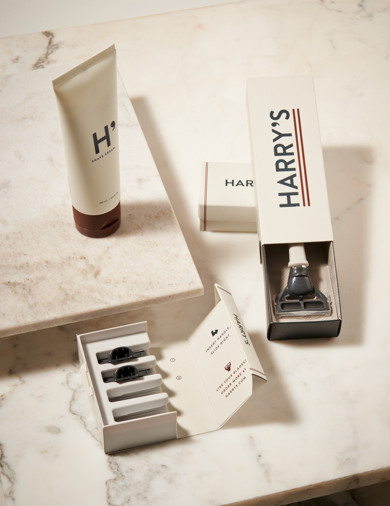 Harry's shaving kit displayed on marble surface with cream tube, razor blades, and branded boxes.