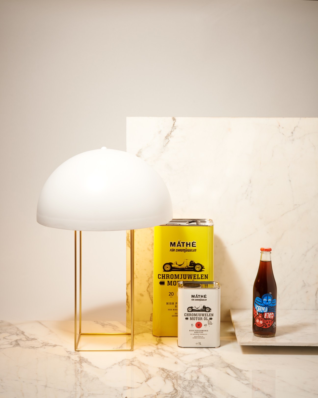 White dome lamp, yellow Chromjuwelen motor oil canister and bottle of Karma Cola soda on marble surface.