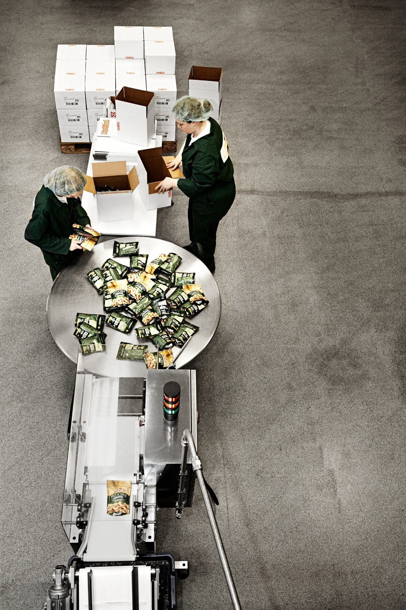 Overhead view of workers in hairnets and green jackets packing Nutisal nut boxes on a production line.