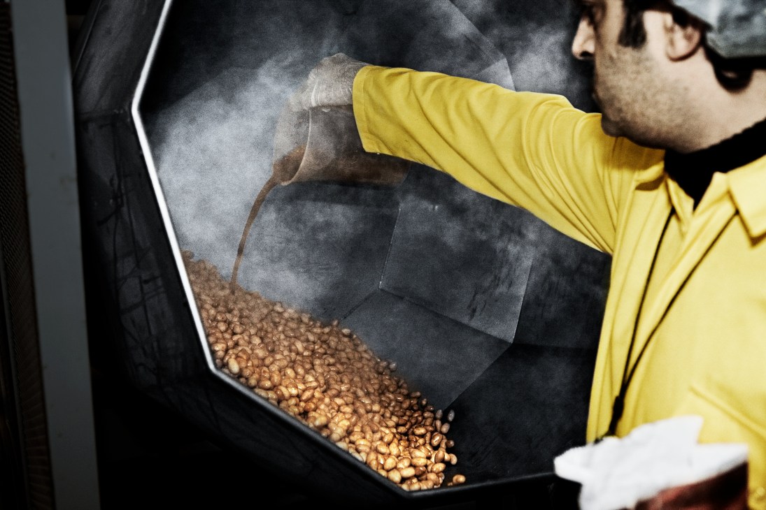 Worker in yellow gloves spraying barbecue flavoring onto pistachios in a large industrial container.