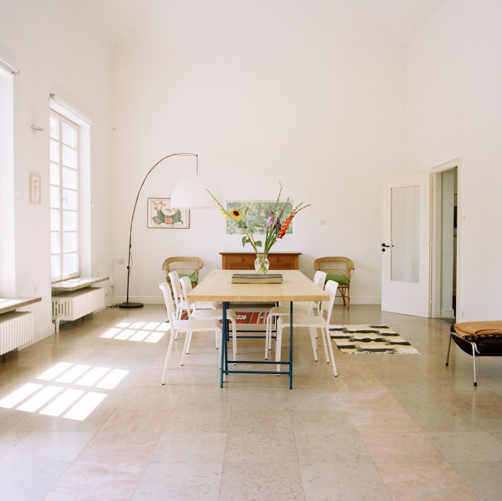 Bright dining room with double-height ceilings, wooden table, woven chairs, and large windows letting in natural light.