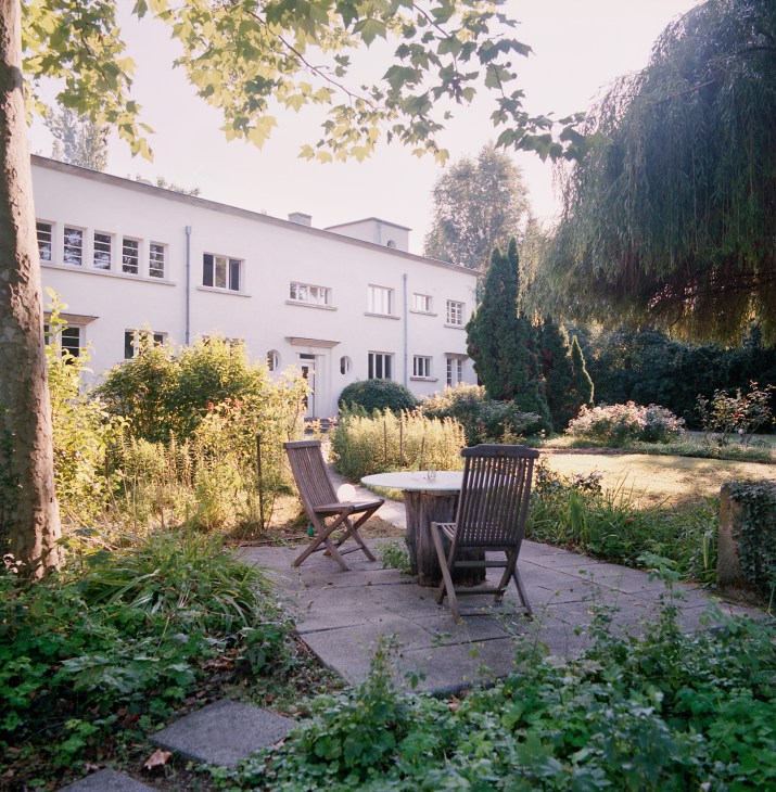 Paved garden seating area with wooden chairs and table, surrounded by manicured hedges and overgrown vegetation beneath overhanging trees at Villa St Martin.