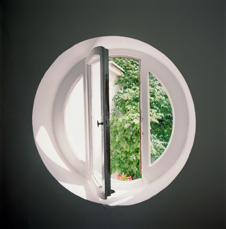Circular white window with dark frame opening onto green foliage and garden beyond.