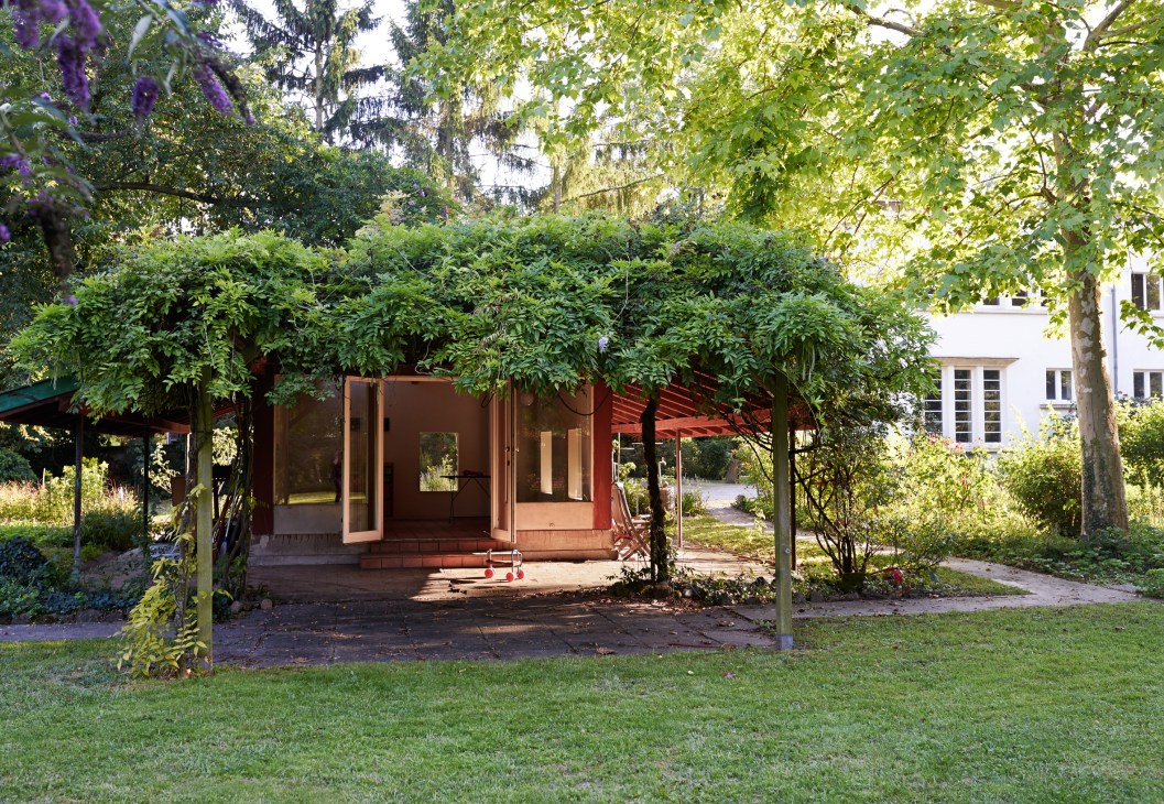 Red-brown modernist pavilion with large windows nestled under mature trees in a garden setting.