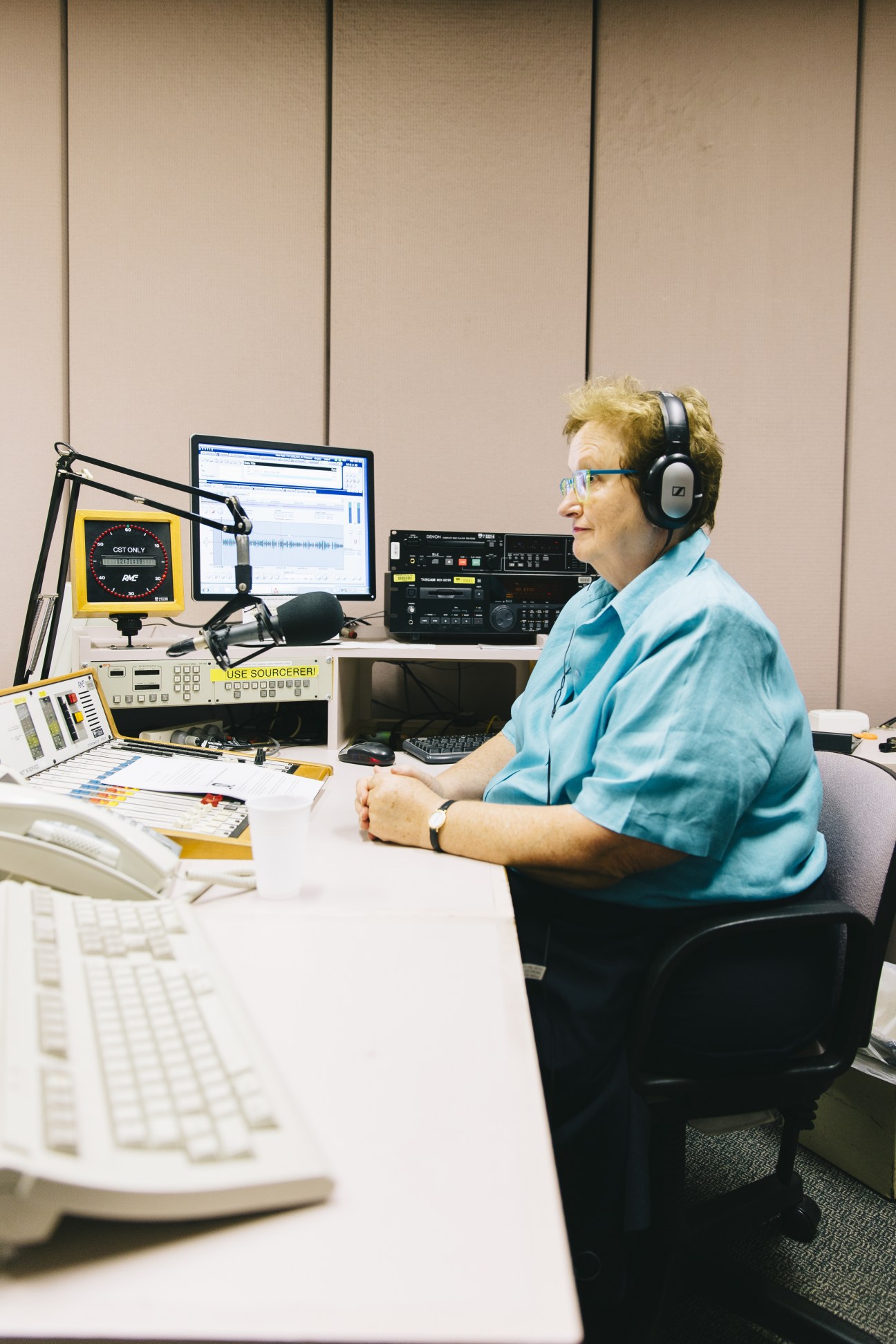 Amanda Vanstone at a radio studio desk wearing headphones and a light blue shirt, with microphone and editing equipment visible.