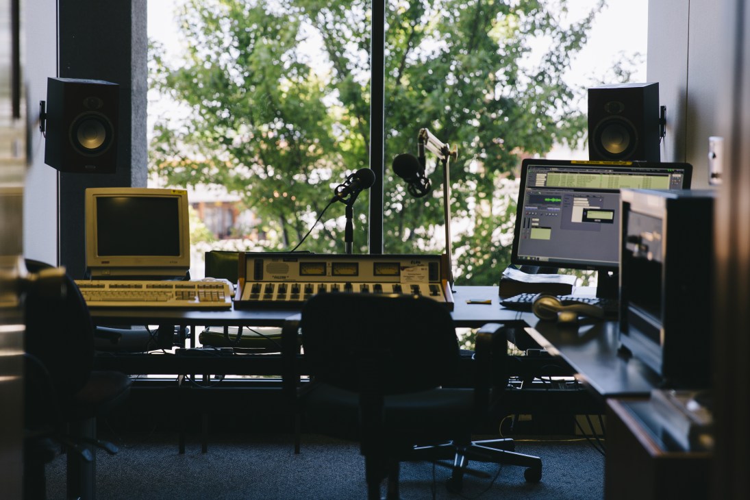 Radio studio equipment including mixing console, microphone, speakers and computer monitors with trees visible through large windows.