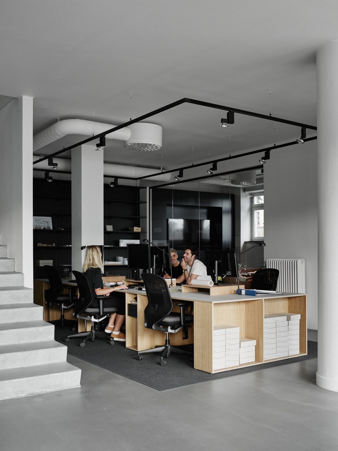 Open-plan office with wooden desks, black chairs, exposed ceiling with track lighting, and white columns