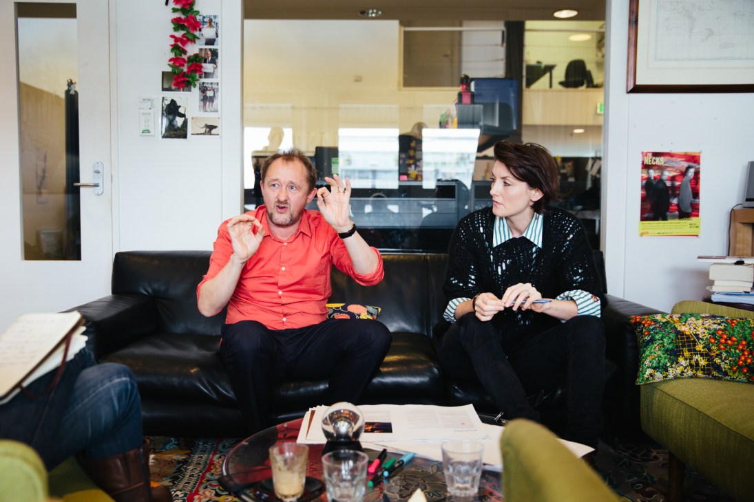 Andrew Upton in a red shirt gestures while speaking with a woman in a black blazer seated on a black sofa in an office.