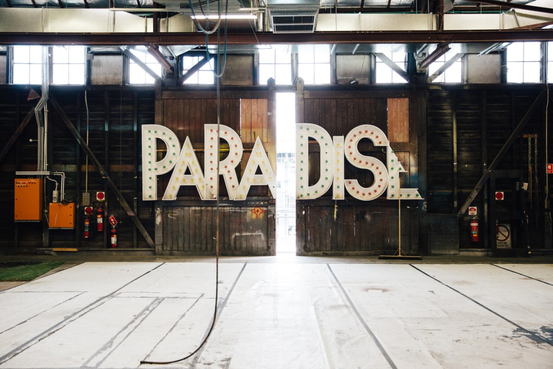 Large illuminated 'PRADA DS' sign in an industrial warehouse with high windows and wooden doors.