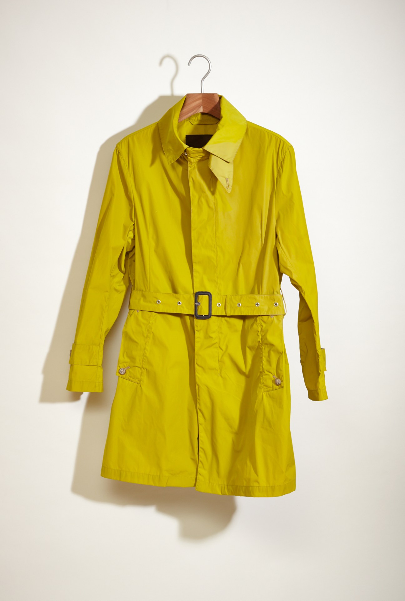 Bright yellow nylon Mackintosh raincoat with belt and polished buttons on wooden hanger against white background.