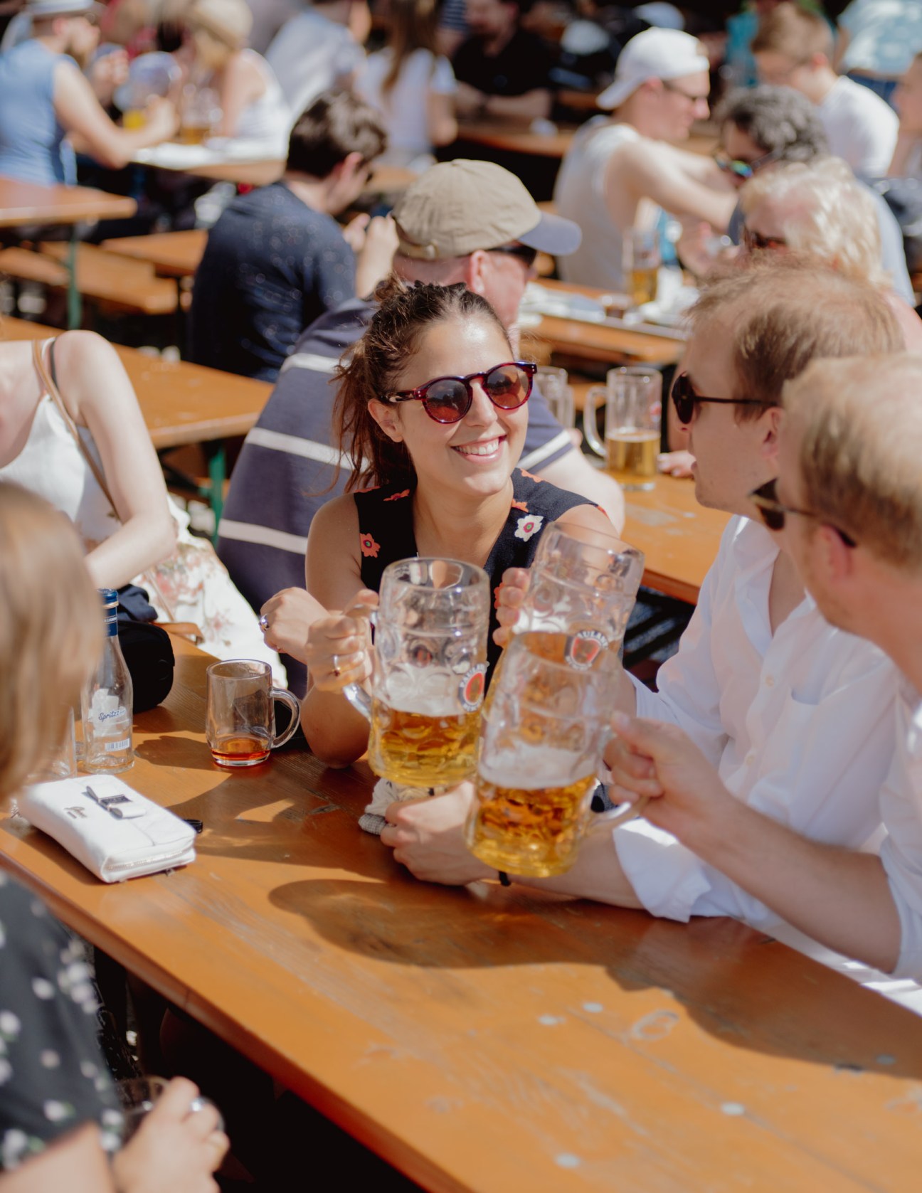 Woman in sunglasses smiling at a beer garden table with beer glasses and other guests around her.