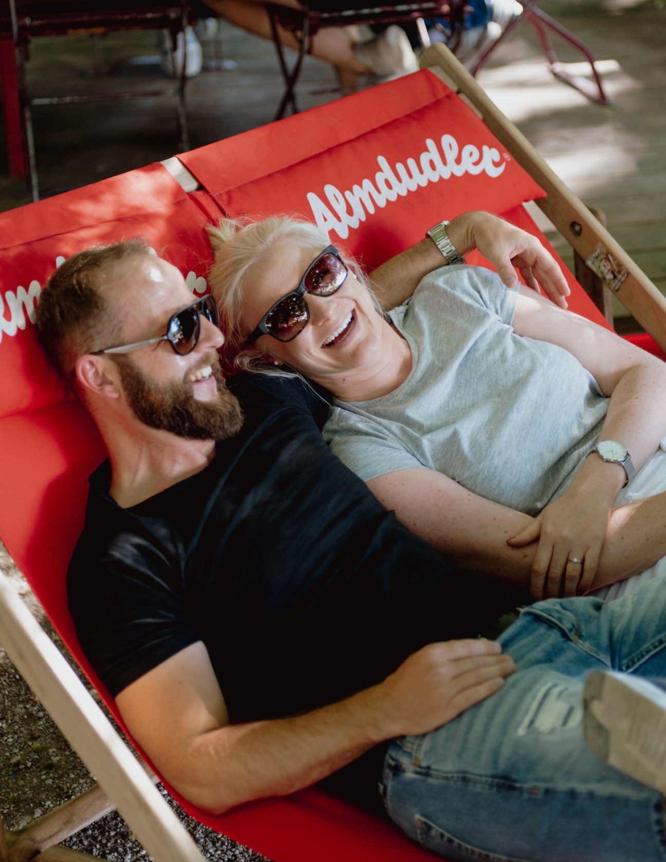 Two people wearing sunglasses smile while relaxing on a red beer garden bench.