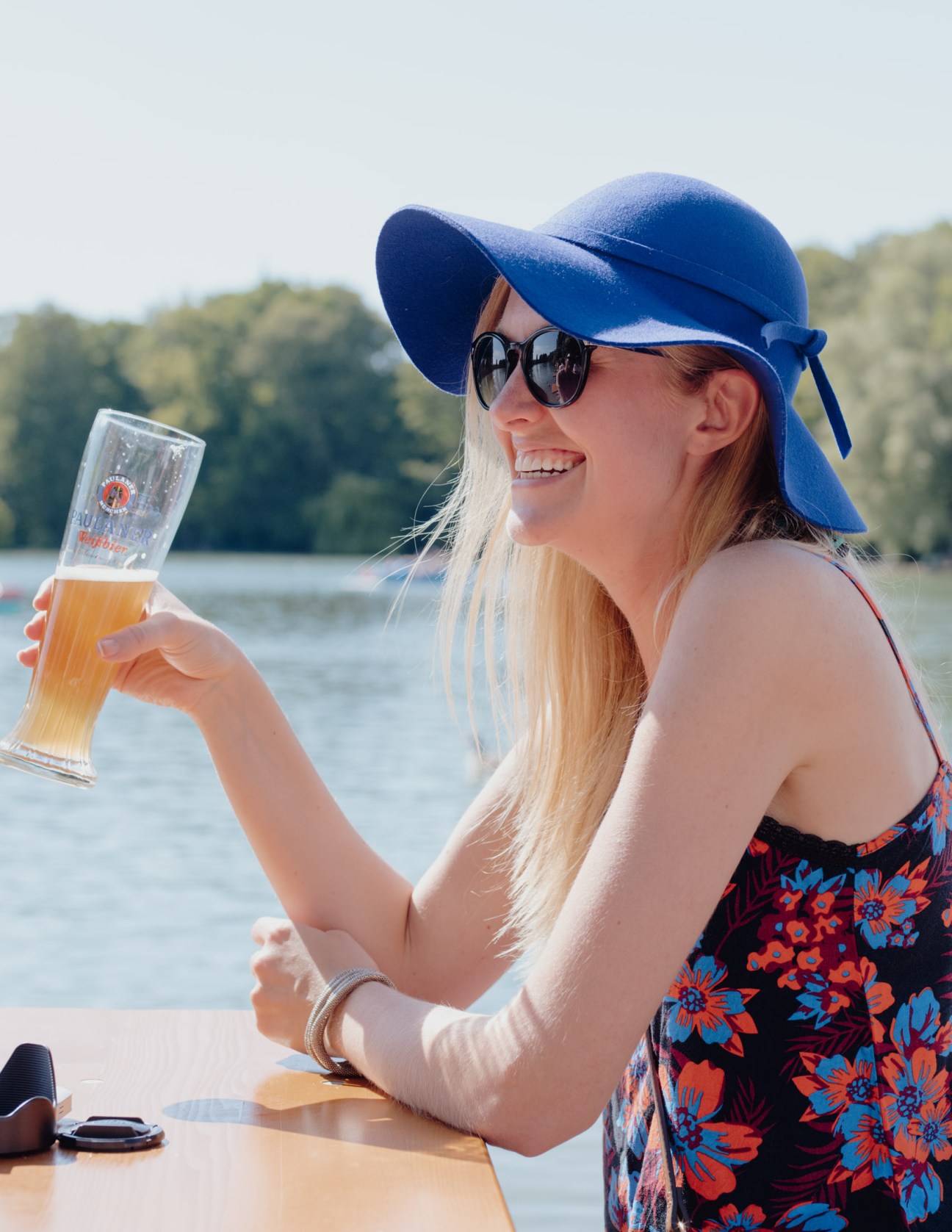 Woman in blue sun hat and sunglasses holding a beer glass by a lake