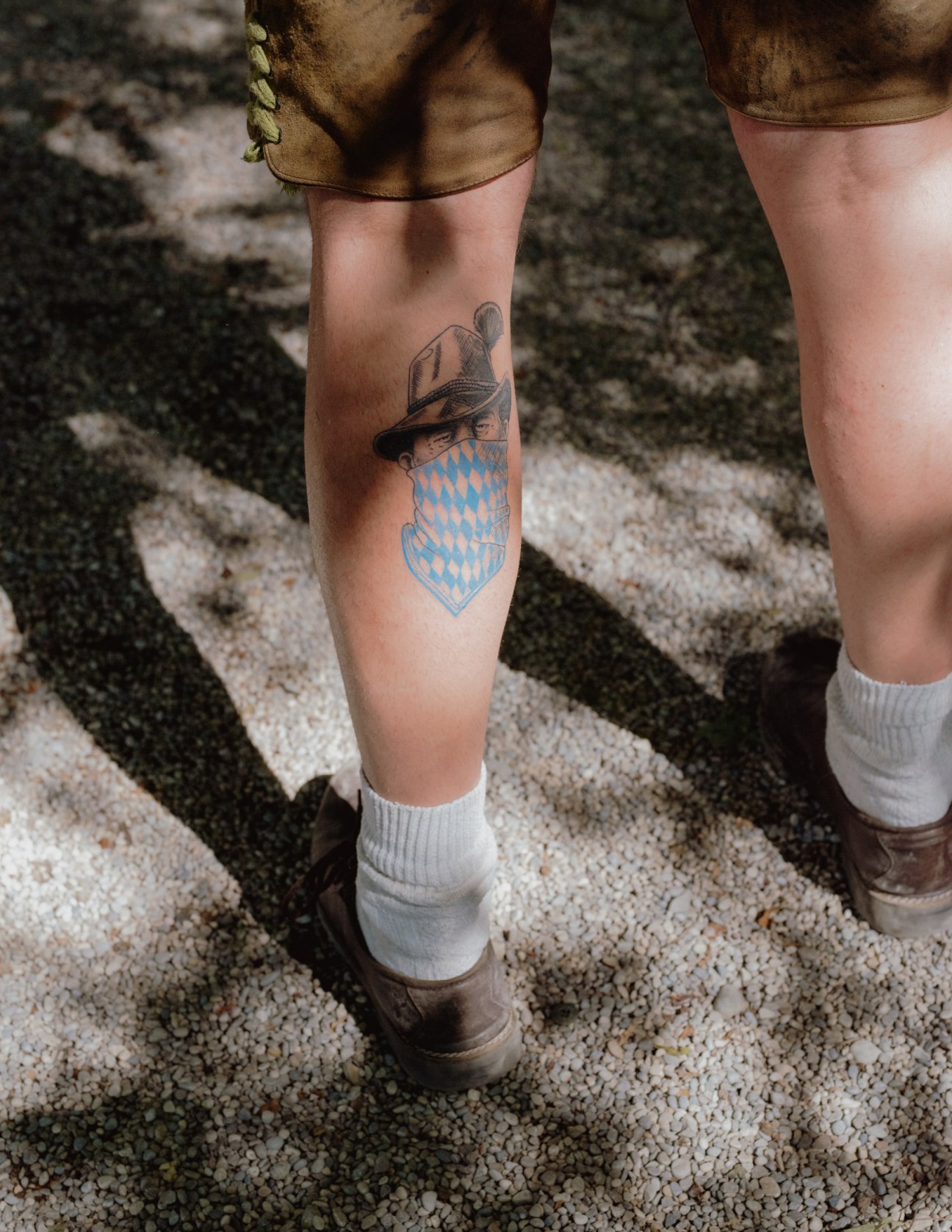 Person wearing traditional Lederhosen and white socks displays a tattoo of a Bavarian hat and blue diamond pattern on their leg.