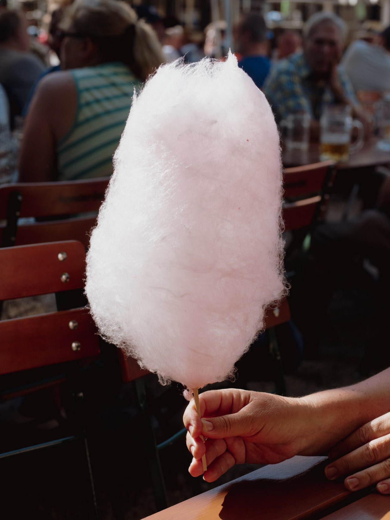 Cotton candy held in hand at a crowded beer garden with wooden benches in background.
