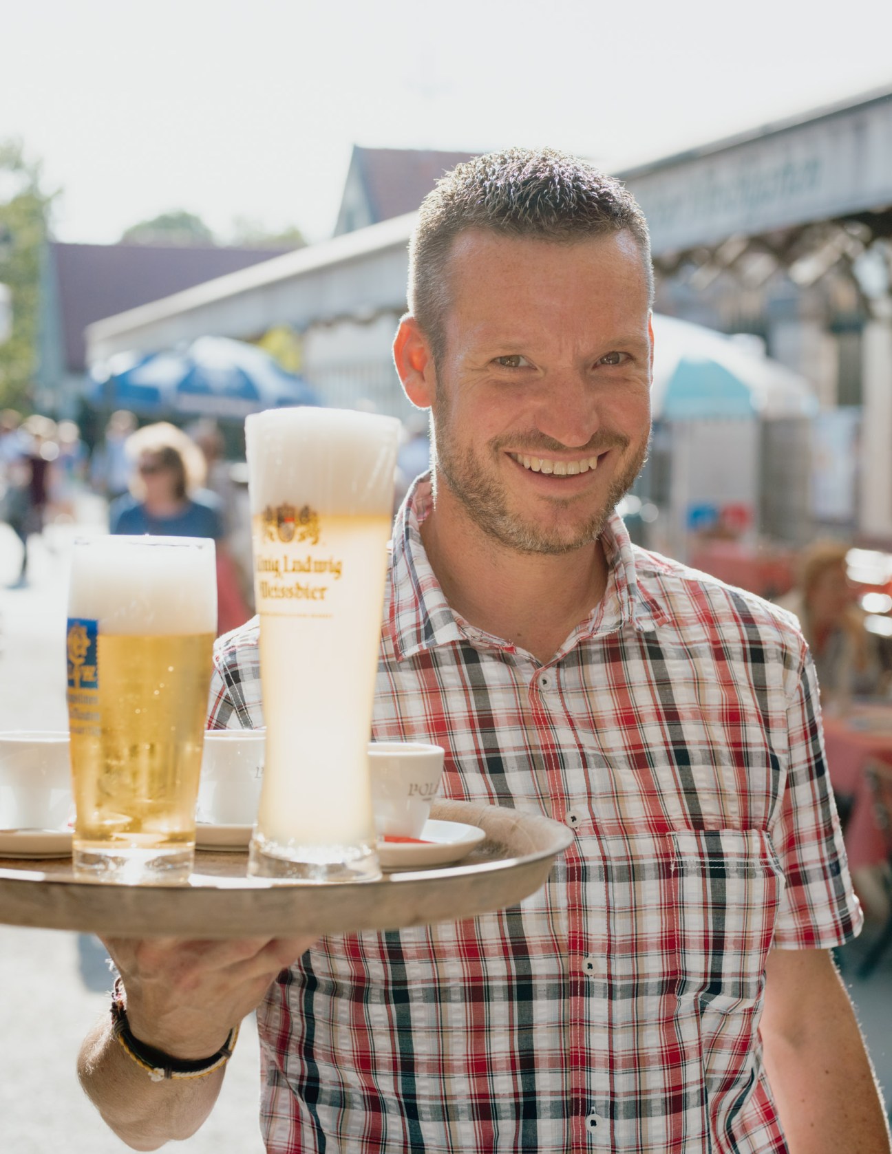 A smiling waiter in traditional Bavarian attire carries a tray with beer glasses at a Munich beer garden.